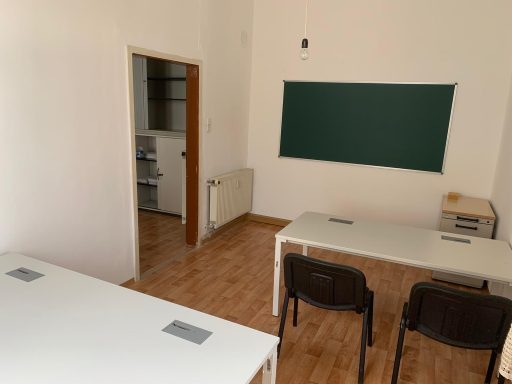 Leeres Klassenzimmer mit zwei Tischen, Stühlen und einer Tafel an der Wand.